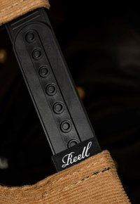 Schwarzer Kunststoffriemen mit runden Knöpfen und einer strukturierten Oberfläche, befestigt an einem beigen Stoffriemen mit einem weißen "Reell"-Logo.