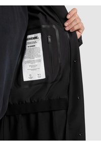 Giacca nera con una texture liscia, dotata di una tasca interna con zip. L'etichetta interna include istruzioni per la cura e informazioni sul marchio.