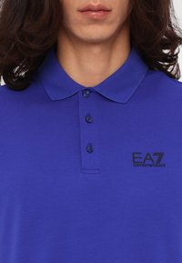 Mėlyna polo marškinė su stojamuoju kaklu, trimis mygtukais ir juodu EA7 Emporio Armani logotipu. Lygus audinys, trumpomis rankovėmis.