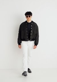 Άνδρας με μαύρο μπουφάν μπόμερ, μαύρο πουκάμισο, άσπρο παντελόνι, μαύρα παπούτσια και γυαλιά ηλίου περπατά σε απλό λευκό φόντο.