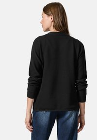 Schwarzer gerippter Pullover mit rundem Ausschnitt, langen Ärmeln und umgeschlagenen Handgelenken; die Rückansicht zeigt eine lockere Passform und strukturierte Stoffoberfläche.