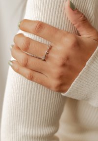 Bague en argent en forme de vague avec deux accents arrondis, portée sur une main à la peau claire. La main est manucurée avec du vernis à ongles vert, reposant sur un pull crème à côtes.