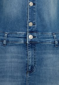 Macacão de ganga em azul médio, com botões de pressão prateados na frente, cintura ajustada e textura de tecido suave com ligeira descoloração.