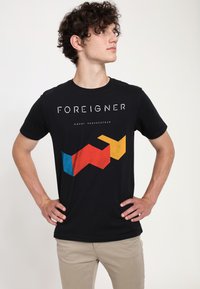 Černé bavlněné tričko se znakem "FOREIGNER" v bílé barvě a barevnými geometrickými tvary v červené, modré a žluté na spodní části předního dílu.