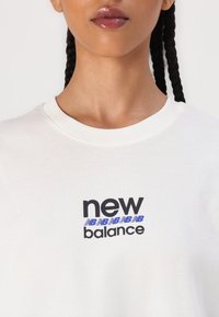 Balts kokvilnas t-krekls ar melnu un zilu "new balance" logo, kas izveidots modernā fontā priekšpusē. Gluda tekstūra, apaļa apkakle, īsas piedurknes.