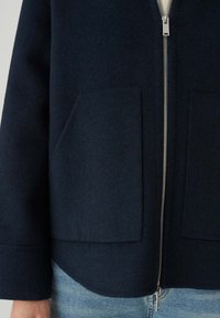 Giacca in lana blu navy con zip frontale, grandi tasche applicate e orlo arrotondato. Tessuto liscio dal design pulito e minimalista.