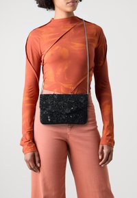 Zwarte met kralen versierde schoudertas met een gestructureerde buitenkant en afsluitbare sluiting, hangend aan een zilveren kettingriem, gedragen over een oranje lange mouwen top.