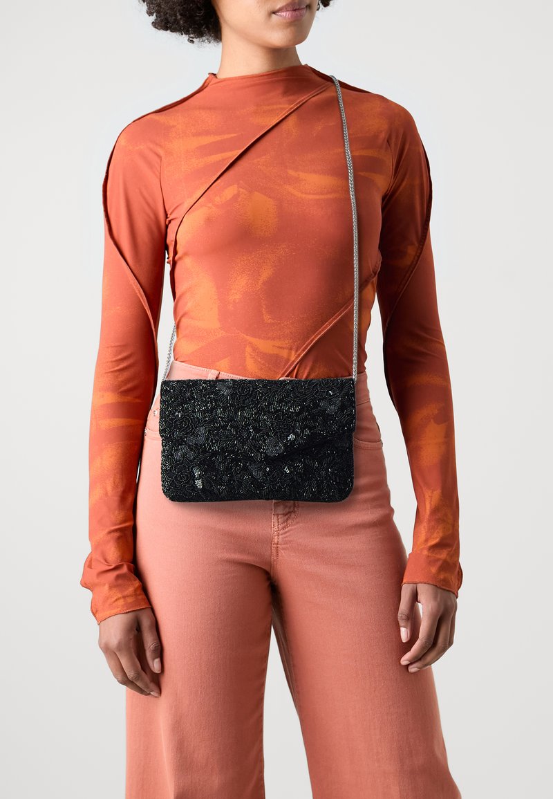 Zwarte met kralen versierde schoudertas met een gestructureerde buitenkant en afsluitbare sluiting, hangend aan een zilveren kettingriem, gedragen over een oranje lange mouwen top.