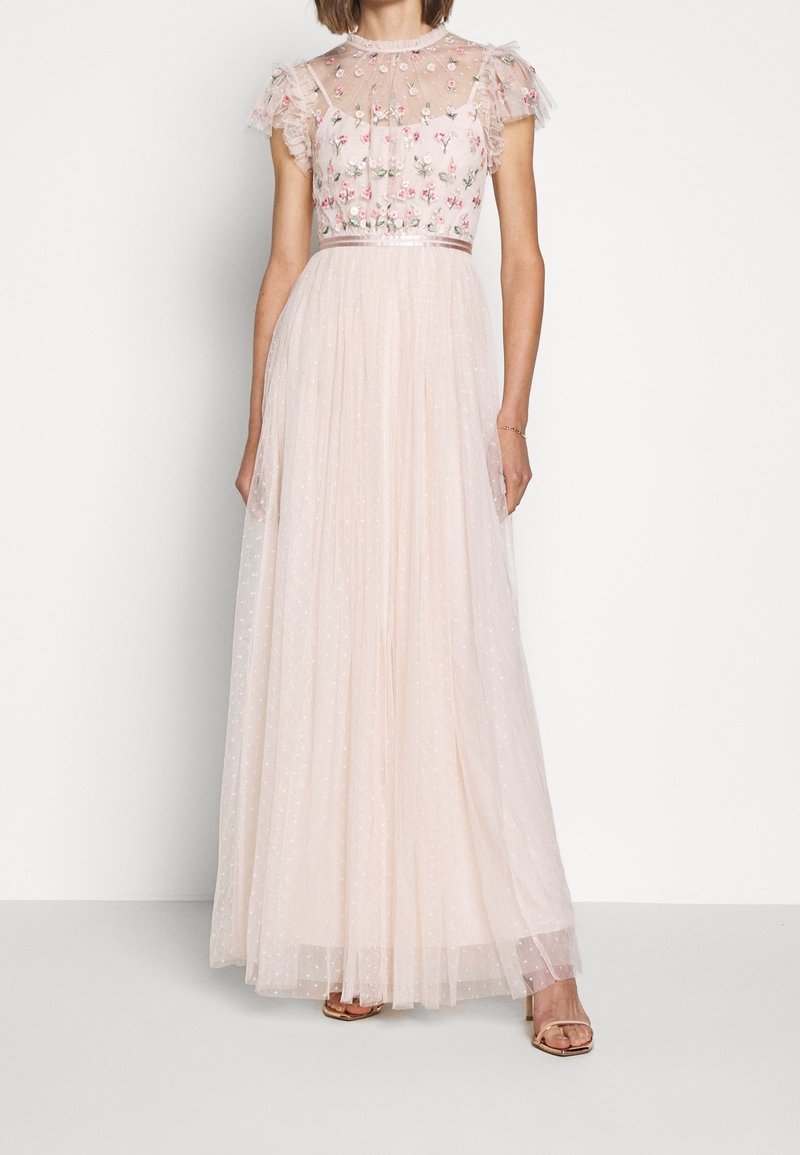 Femme portant une robe longue rose pâle avec un corsage floral brodé transparent et des manches courtes à volants, debout sur fond blanc.