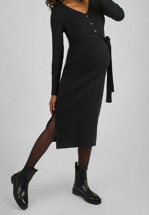 Zwangere vrouw draagt een zwarte midi-jurk met lange mouwen, knopen en een zijsplit, zwarte panty's en zwarte enkellaarsjes, staand op één been.