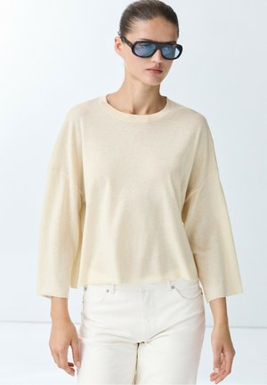 Donna che indossa una maglia beige oversize a maniche lunghe, pantaloni bianchi e grandi occhiali da sole neri con lenti blu, in piedi davanti a uno sfondo semplice.