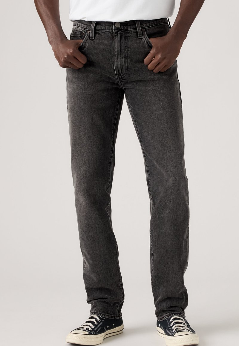 Homme portant un jean slim noir et des baskets basses noires avec des lacets et semelles blanches, les mains reposant près des poches avant.