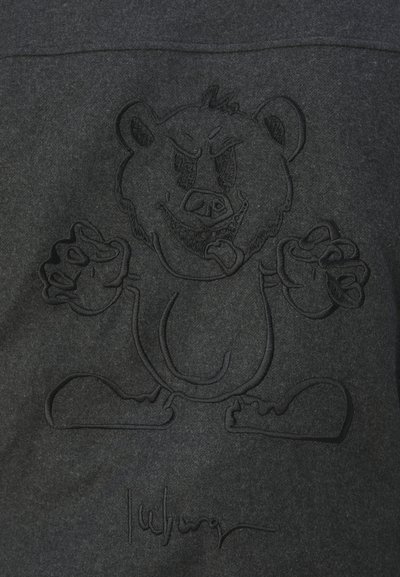 Sudadera negra con un gráfico de oso en relieve que presenta características faciales y manos exageradas, junto a una firma en la parte inferior.