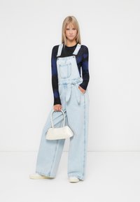 Lichtblauwe denim overall met wijde pijpen, verstelbare bandjes, voorzak en een strikdetail in de taille, gecombineerd met een witte handtas en schoenen.