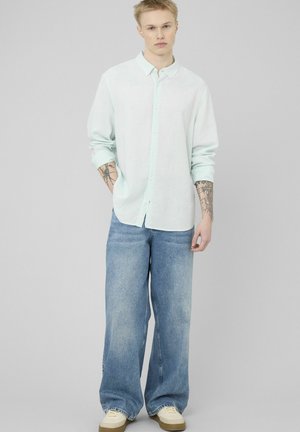 Jeune homme avec des tatouages sur les bras, portant une chemise à boutons vert menthe claire, un jean bleu à jambes larges, et des baskets crème, debout devant un fond uni.