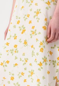 Katoenen jurk met een witte gematteerde stof en een bloemenpatroon van gele en groene bloemen. De jurk heeft een losse pasvorm en is tot op de knie.