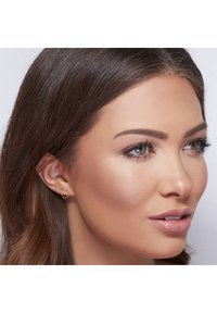 Roze gouden hartvormige studs; dun, delicaat ontwerp; gladde textuur; gedragen in het linker oor; aangevuld met natuurlijke make-up en donker haar.