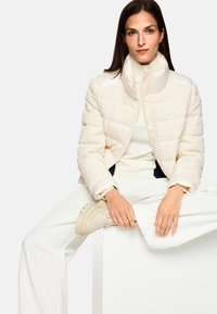 Steppjacke in Creme mit strukturierter Stoffoptik, hohem Kragen und Reißverschluss. Kombiniert mit cremefarbenen Hosen und robusten beigen Sportschuhen.