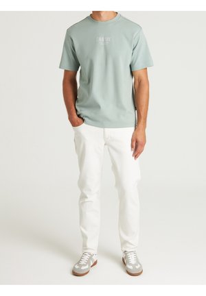 Hellgrünes Baumwoll-T-Shirt mit Rundhalsausschnitt und Frontlogo. Kombiniert mit weißen Jeans und grauen Sneakers mit braunen Sohlen.