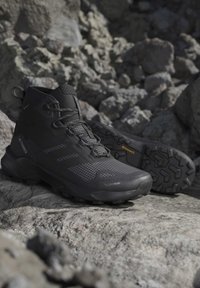 Black high-top hiking shoes with textured soles on rocky terrain, featuring grip patterns and branded details visible on sides and tongue.