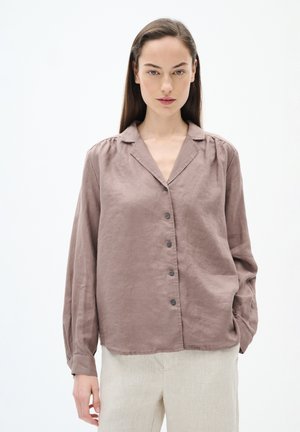 Brown linen long-sleeve shirt with subtle pleats, notched collar, and six dark buttons; paired with light beige linen trousers.