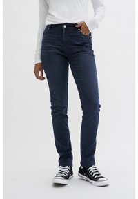 Jeans de mezclilla azul oscuro con un corte ajustado, diseño de cinco bolsillos y cierre frontal con botón, combinados con zapatillas negras y blancas.
