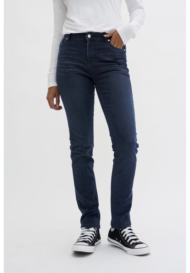 Jeans de mezclilla azul oscuro con un corte ajustado, diseño de cinco bolsillos y cierre frontal con botón, combinados con zapatillas negras y blancas.