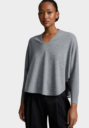 Femme portant un haut ample gris à col en V et manches longues et un pantalon noir, debout devant un fond uni gris clair.