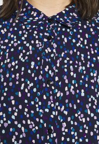 Gros plan d'une chemise à col bleu foncé avec des boutons noirs et un motif dispersé de points irréguliers de couleur violet clair, bleu et blanc.
