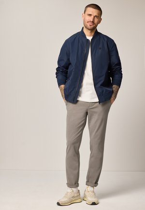 Mężczyzna stoi z rękami w kieszeniach, ubrany w granatową kurtkę, białą koszulkę, szare spodnie, beżowe trampki oraz widoczne tatuaże na ramionach na jednolitym tle.