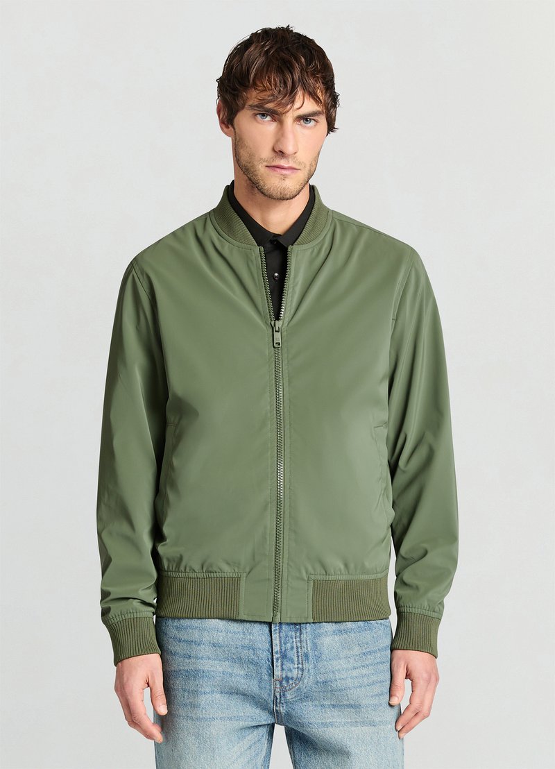Olive green bomber jacket, lightweight, zip-up front, ribbed collar and cuffs, smooth texture, paired with blue denim jeans.