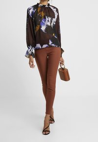 Schwarze Bluse mit mehrfarbigem Blumenmuster, Rüschenausschnitt und plissiertem Stoff. Braune Slim-Fit-Hose und eine kleine, strukturierte braune Handtasche.