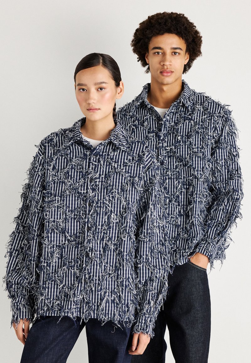 Aplūsto, auduma krekli ar tumši zilu un baltu svītru rakstu, ar pārspīlētu izmēru, teksturētām pavedienu detaļām un garām piedurknēm, saskaņoti ar tumšām džinsēm.