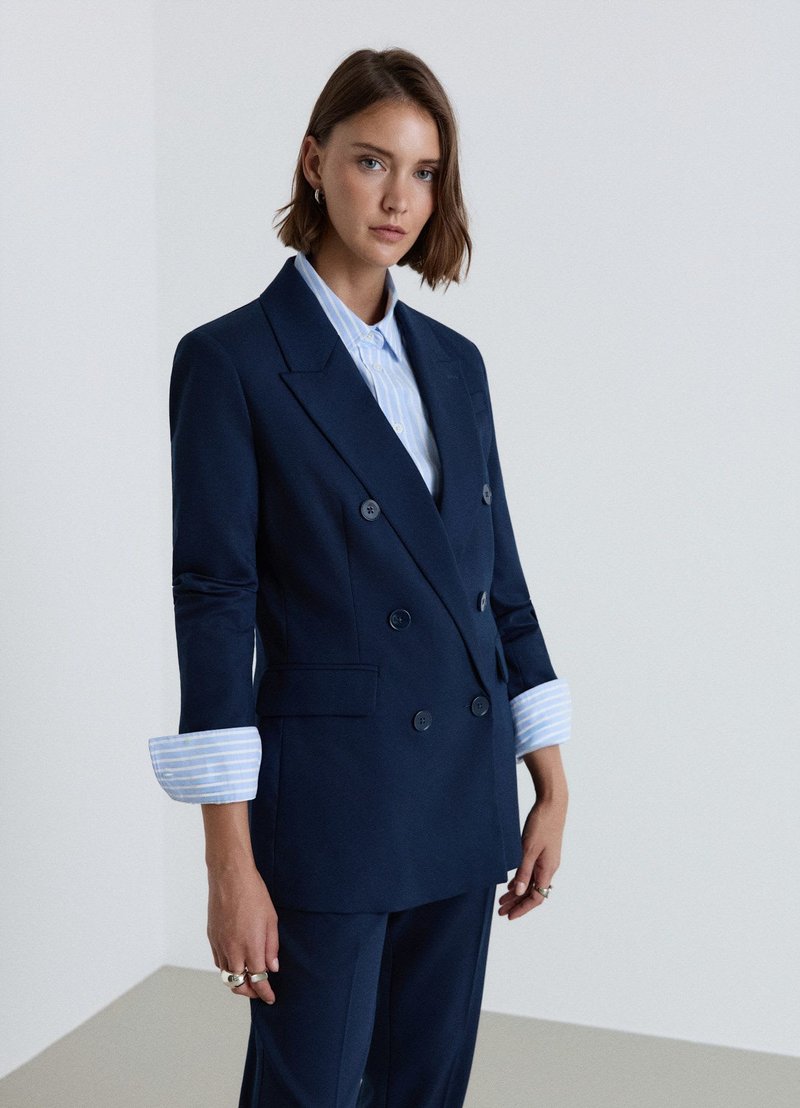 Woman wearing a navy blue double-breasted blazer with rolled-up sleeves over a light blue striped shirt, standing against a plain white background.