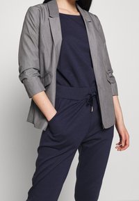 Grå skräddarsydd blazer med en slät textur, notch-lapel och uppkavlade ärmar, kombinerad med en marinblå topp och matchande joggerbyxor med dragsko.