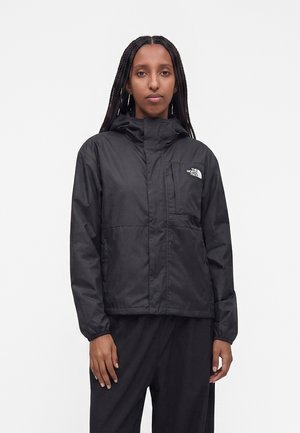 Personne aux longs cheveux tressés portant une veste noire à capuche et un pantalon avec le logo The North Face sur la poche poitrine.