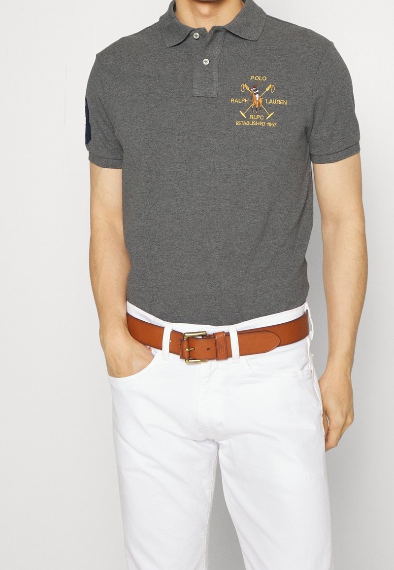Homme portant un polo gris foncé avec le logo jaune Polo Ralph Lauren, un pantalon blanc et une ceinture en cuir marron, debout avec une main dans la poche.
