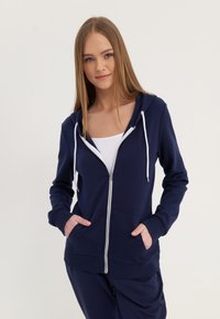 Tamno plava zip-up dukserica napravljena od mekanog materijala s kapuljačom, podesivim šniralicama i prednjim džepovima. Sadrži rebraste manžete i porub.