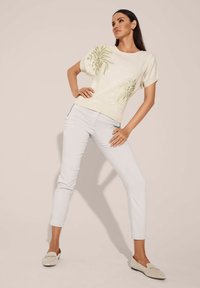 Lichtbeige top met korte mouwen en groene bloem borduursels, gecombineerd met aansluitende witte broeken en lichtgekleurde schoenen. Glad textuur overal.