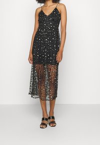 Robe noire avec un motif léopard, dotée d'un corsage ajusté, de bretelles spaghetti et d'une superposition de jupe transparente. Associée à des sandales à talons noires.