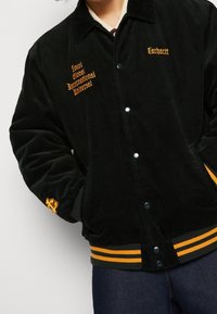 Czarna kurtka sztruksowa z pomarańczowym haftowanym napisem; zatrzaski, ściągacze na mankietach i dole kurtki z żółtymi paskami. Widoczne detale napisu i akcentów.