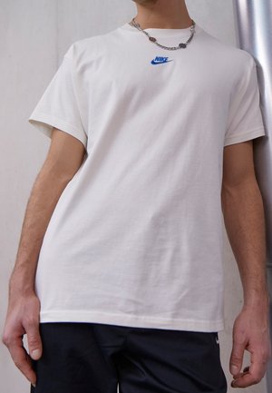 T-shirt blanc en coton à manches courtes, avec un logo Nike bleu sur la poitrine. Porté avec une chaîne argentée.