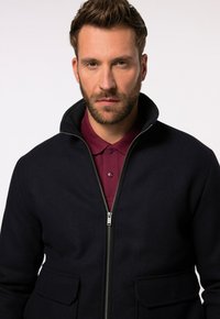 Navy-blauer Zip-Jacke mit hohem Kragen, zwei Fronttaschen und einem weinroten Poloshirt darunter. Strukturierter Stoff, sauberes Design, keine Muster.