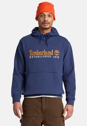 Mann trägt orangefarbene Mütze und dunkelblauen Timberland-Hoodie mit Logo und Text, kombiniert mit brauner Hose, steht vor weißem Hintergrund.