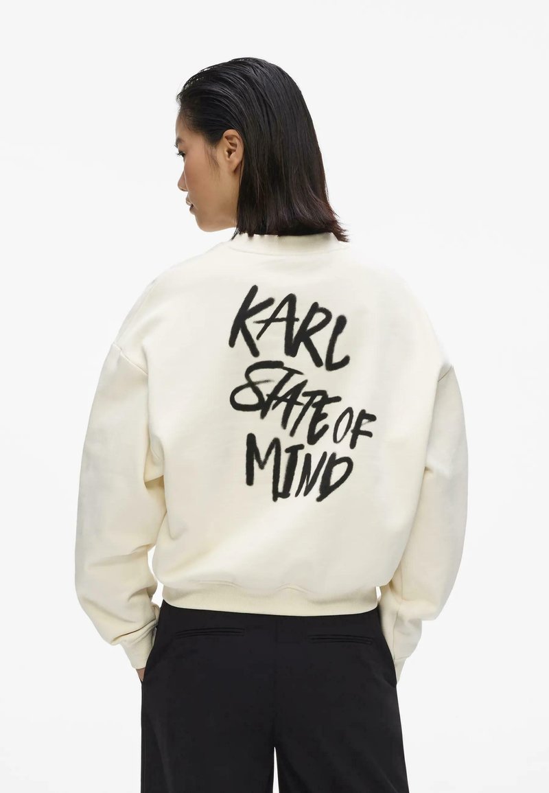 Femme aux cheveux bruns mi-longs portant un sweat crème avec l'inscription « KARL STATE OF MIND » en lettres noires au dos, associée à un pantalon noir.