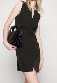 Black sleeveless dress with a V-neck, fitted silhouette, and a front slit, paired with a structured dark handbag featuring cutout handles.