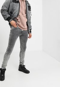 Muž v šedé bomber bundě, růžové mikině, vybledlých šedých skinny džínách a černých vysokých teniskách stojící na bílém pozadí.