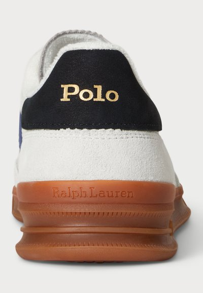 Sneaker branco com calcanhar em camurça azul-marinho, apresentando a marcação dourada "Polo". Sola de borracha laranja com "Ralph Lauren" estampado na lateral.