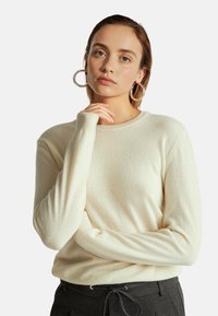 Suéter color crema de material suave, con cuello redondo y puños de canalé. El modelo muestra un corte relajado, combinado con pantalones oscuros.