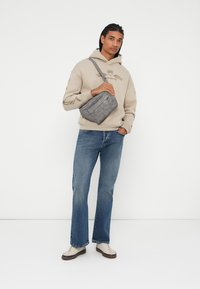 Borsa a tracolla grigia con finitura texturizzata, indossata da una persona con un felpa beige e jeans blu, dotata di una chiusura superiore a zip e due tasche.